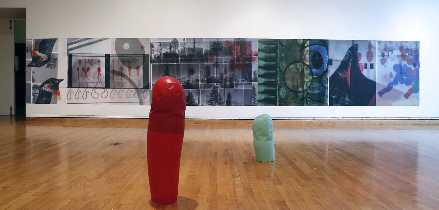 Red abstract sculpture and a smaller green abstract sculpture on a wooden floor in front of an abstract mural.