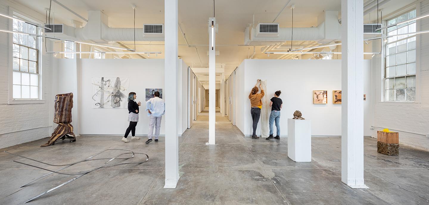 Four students hanging up artwork in a well-lit art studio with cement floors and white columns.