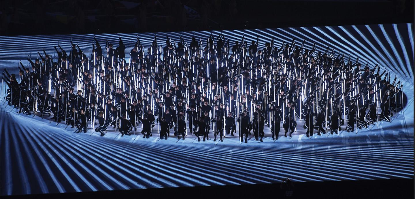 A large group of people huddled into a circle dressed in black holding staffs of light.