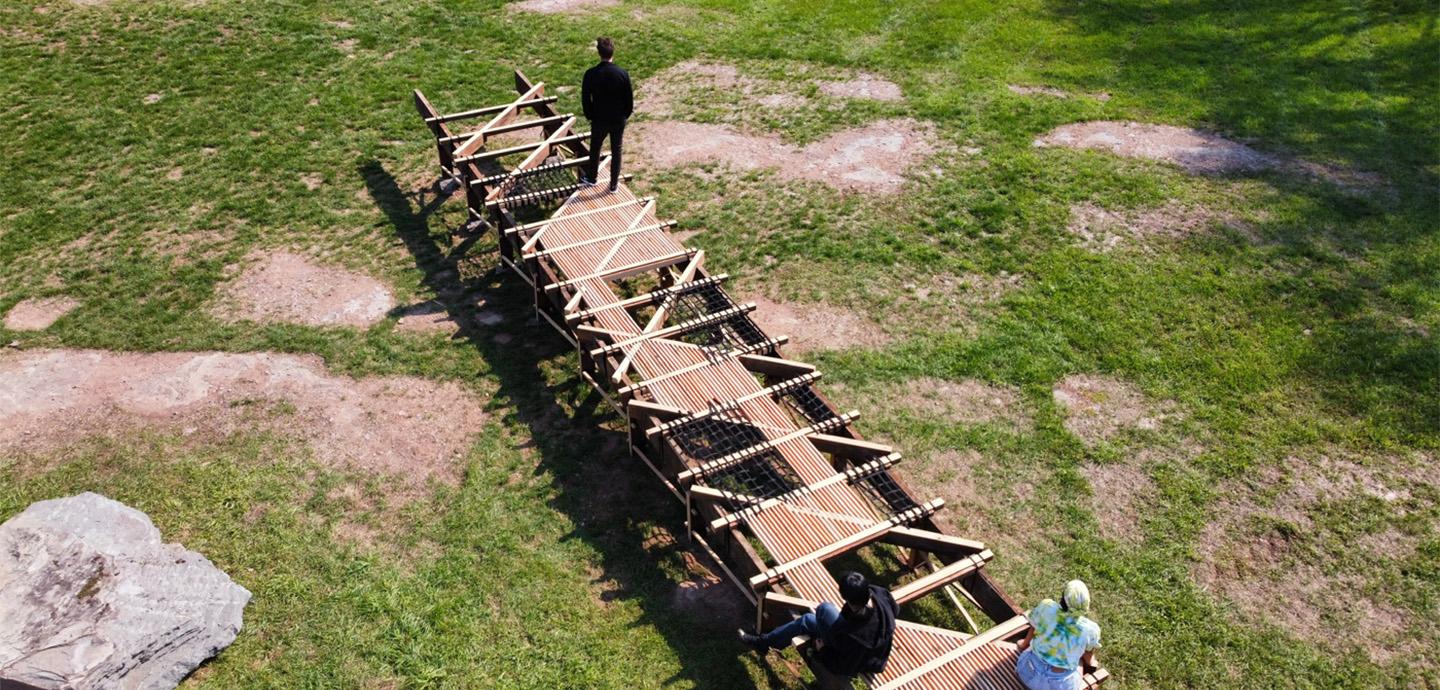 A photo of the wooden boardwalk over grass with 1 person walking on it and 2 people sitting on it