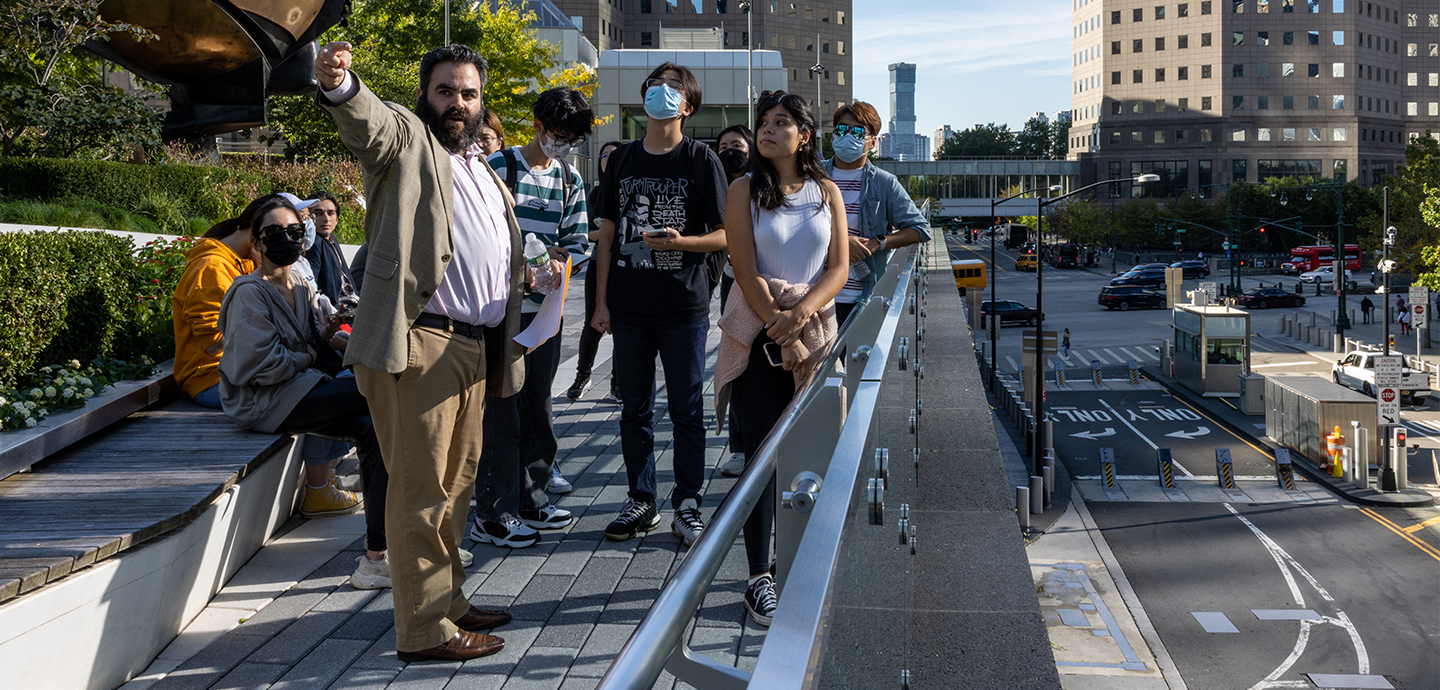 Students gathered on a walkway above an urban street listening to a man in a suit pointing
