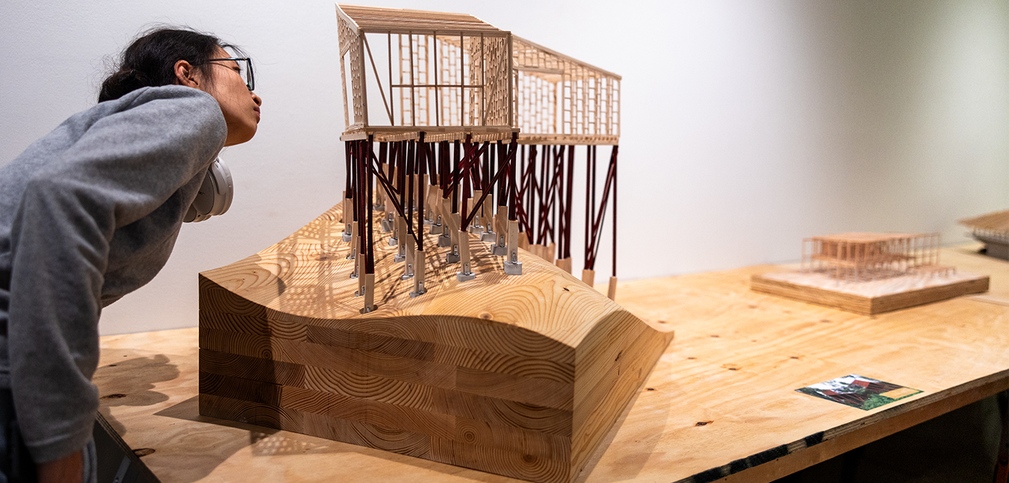 Student inspects a three dimensional structure made from wood and mental components, positioned on top of an undulating wooden block.