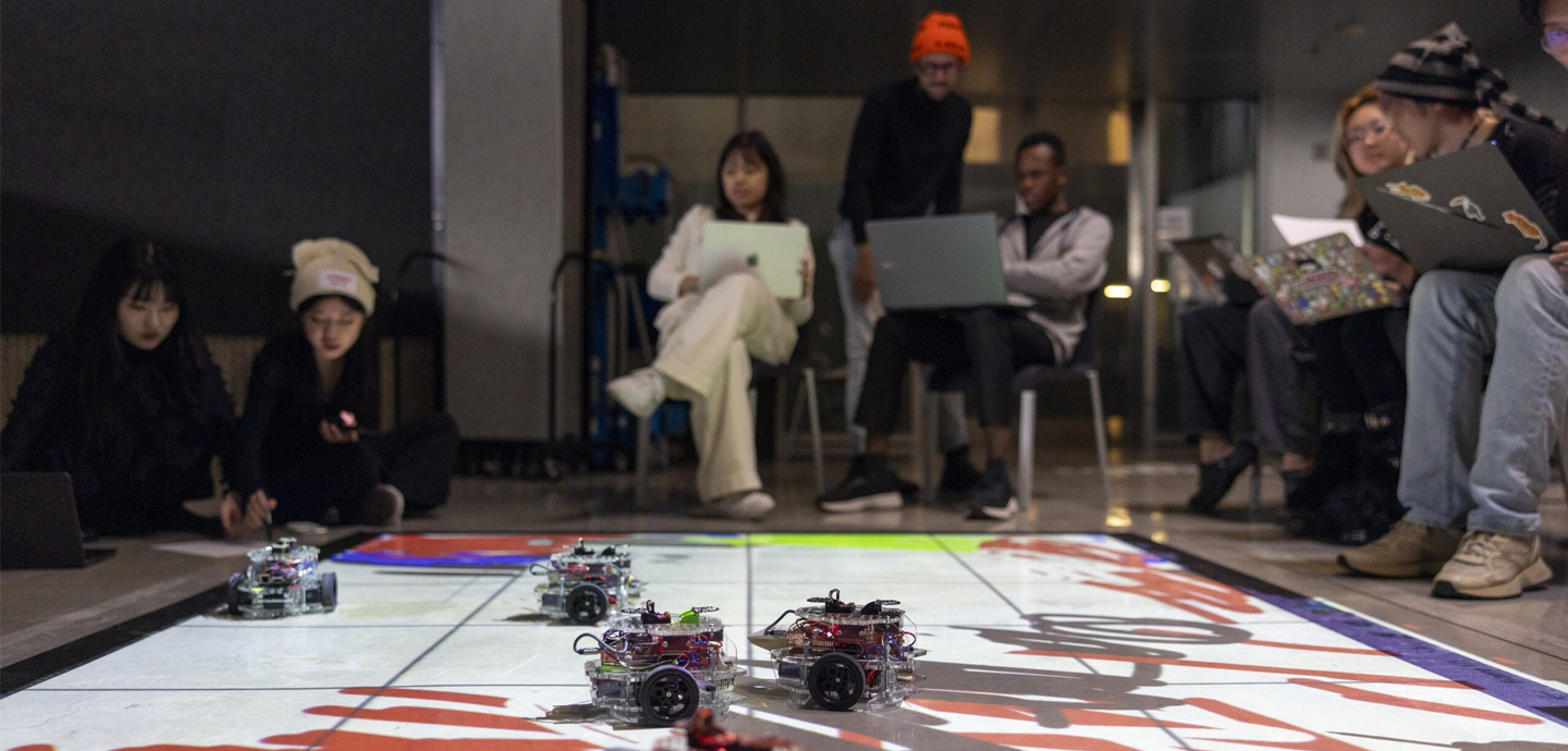 Design Tech students sit around a floor mat that is occupied by five robotic vehicles.