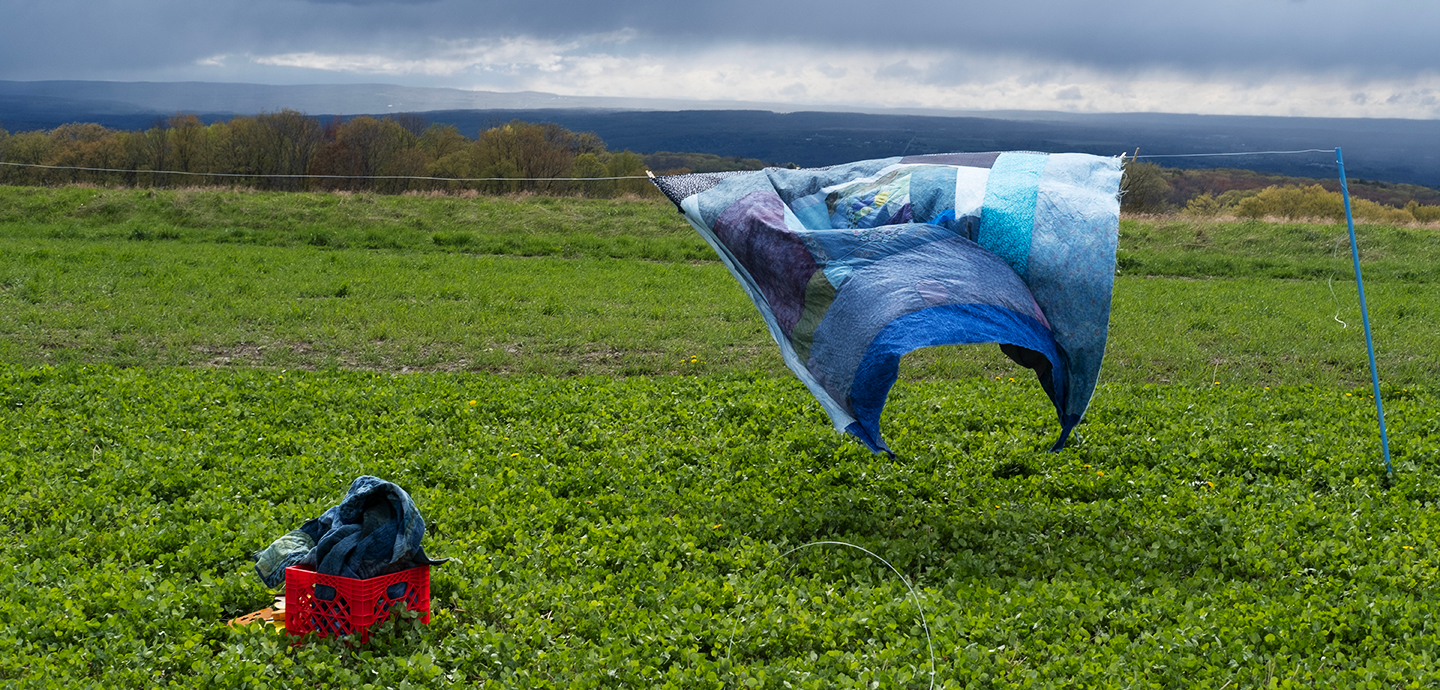 On the left, a red milk crate filled with blue fabric sits in a lush, green field. On the right, a blue  quilt hanging from a clothesline, blowing in the wind with the green ground under it and cloud grey and blue sky behind it. 