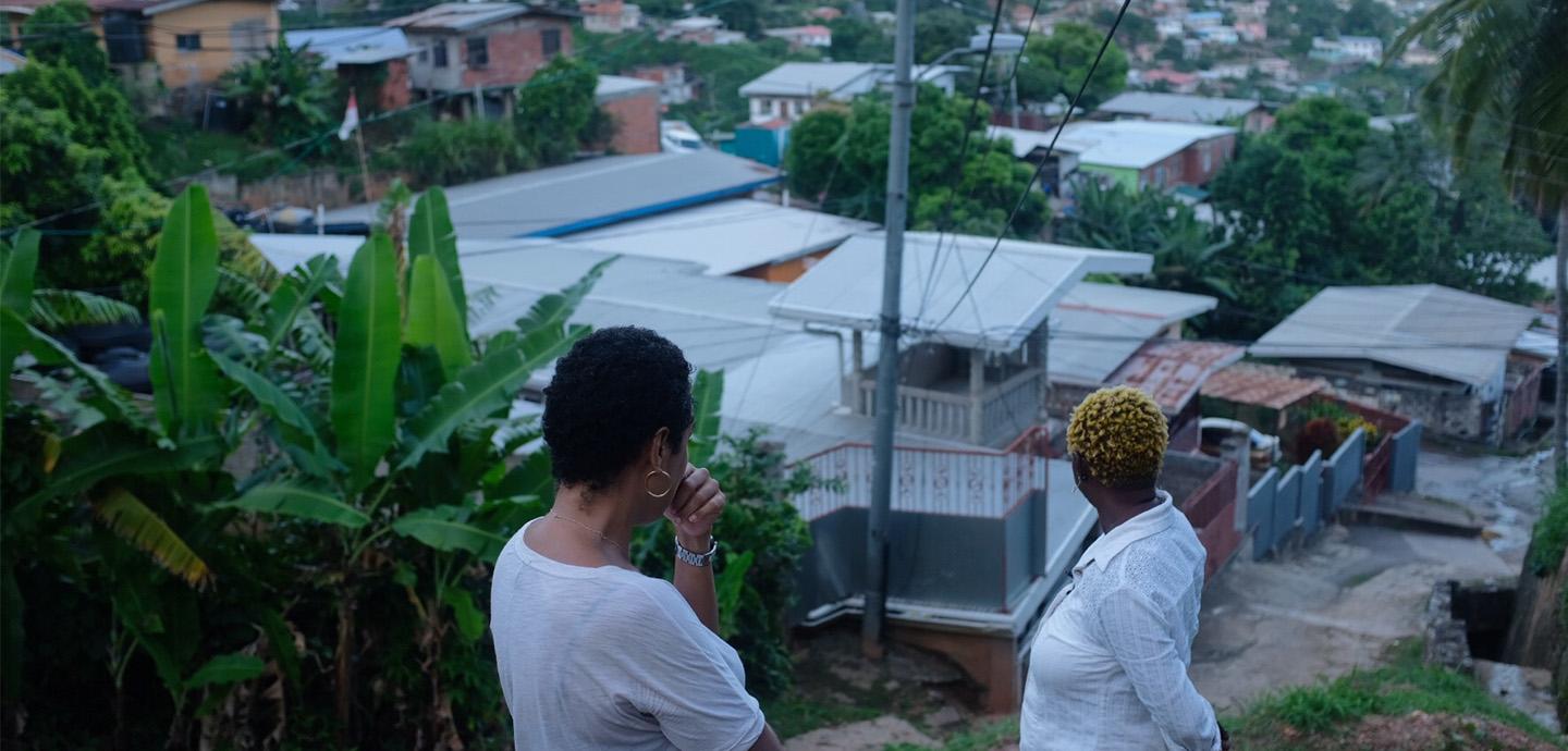 Two people standing side by side, facing towards a landscape consisting of small houses and greenery. 