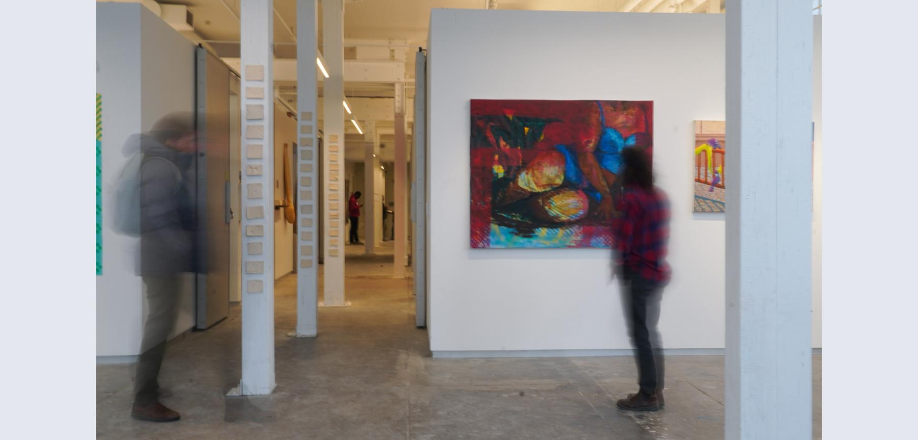 Two people walking in a space looking at paintings on a wall with various white pillars down a hallway.