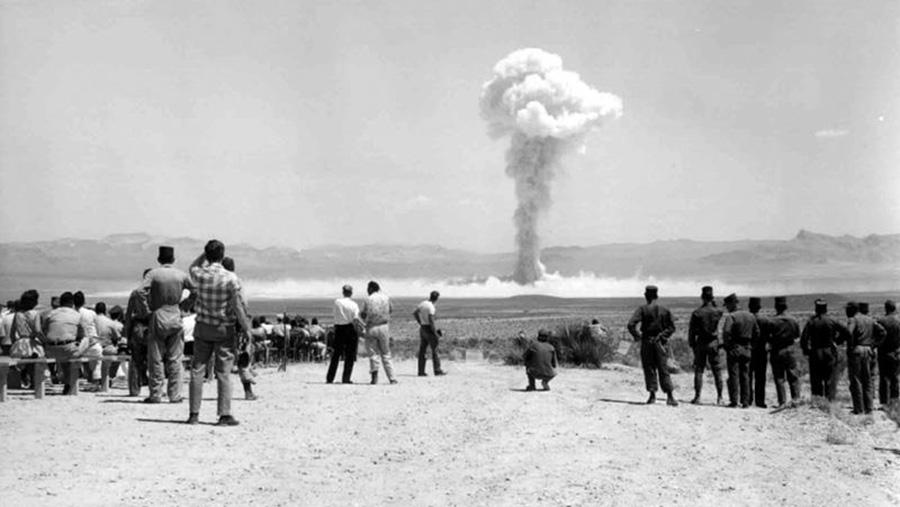 a black and white image of a nuclear cloud in the middle of the desert, the photograph taken behind the people looking on