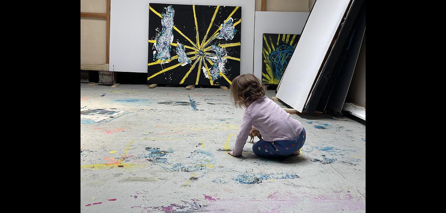 Child playing on a paint covered floor with paintings propped up on the wall.