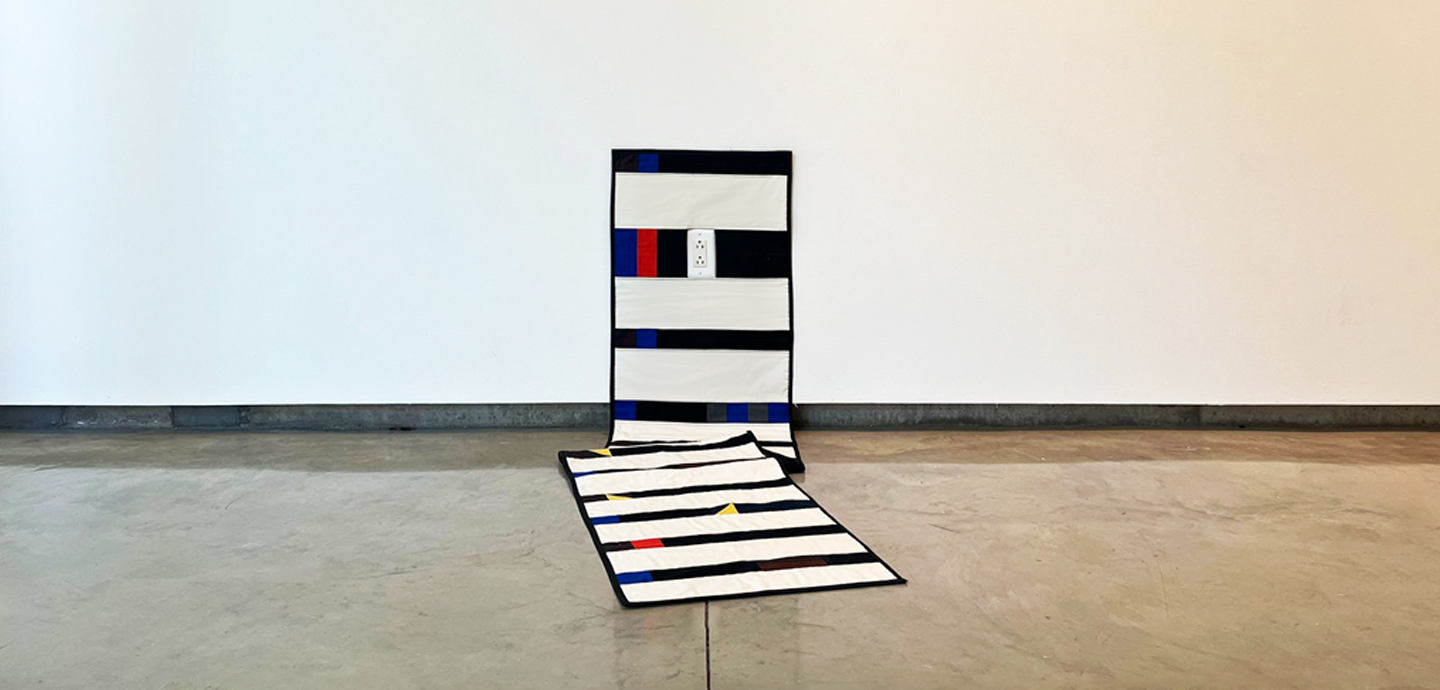 Rectangular quilt with white and black stripes mounted against a white wall, with the tail of the quilt draped on the floor.
