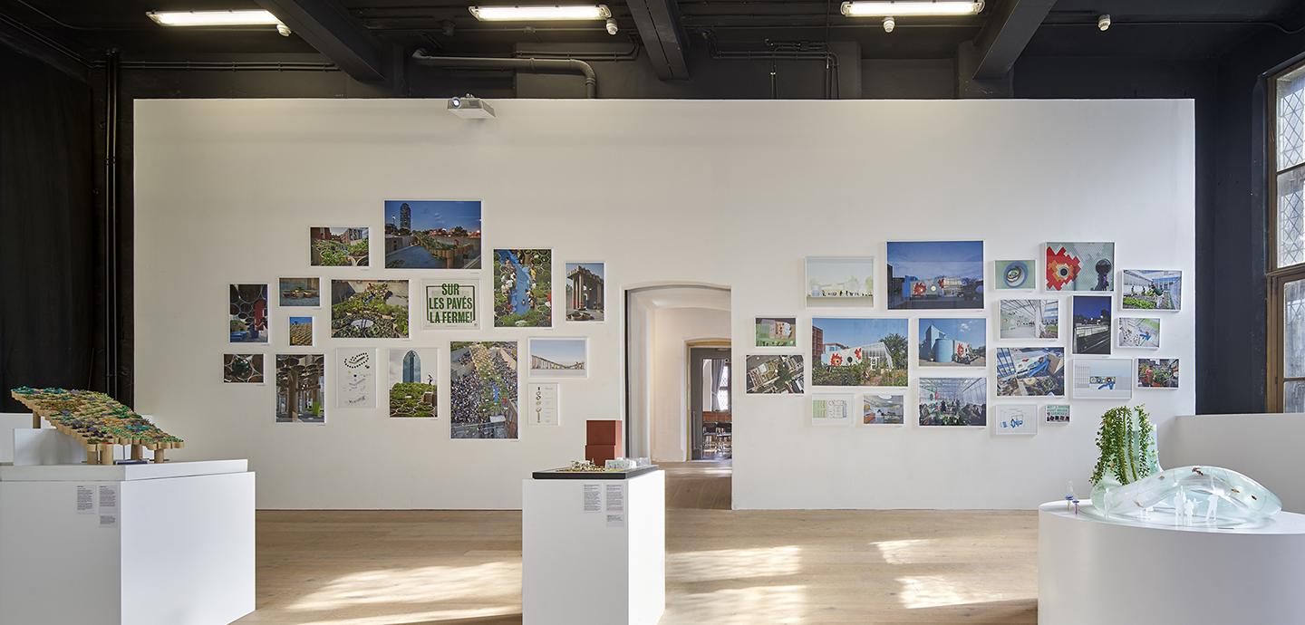 The interior of a modern gallery space. Images are hung on the walls and models are displayed on white podiums varying in size.
