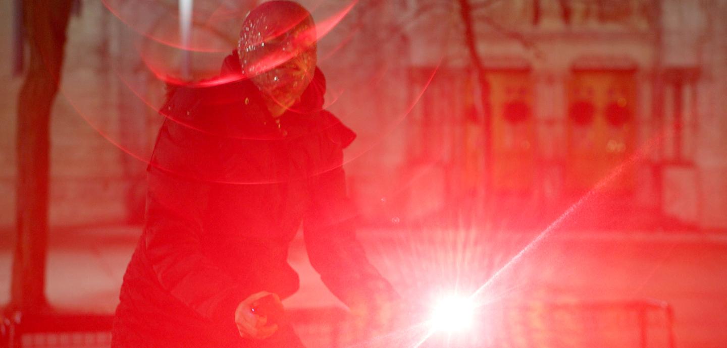 A humanoid figure in a black trench coat stands against a background of a stone building. Overlaid is a bright point of light in the bottom center and a hazy red filter that partially obscures the image.