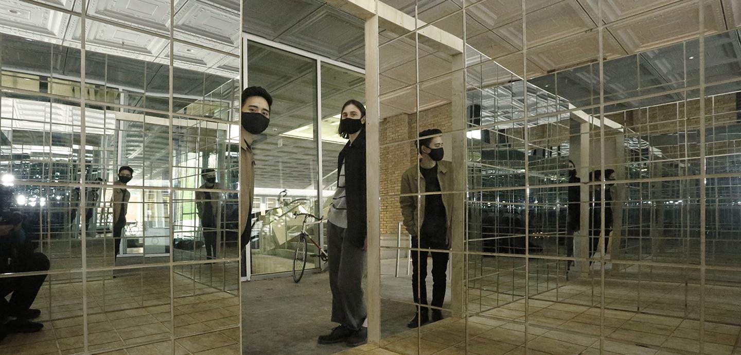 Two people with masks in the doorway of a mirrored box structure.