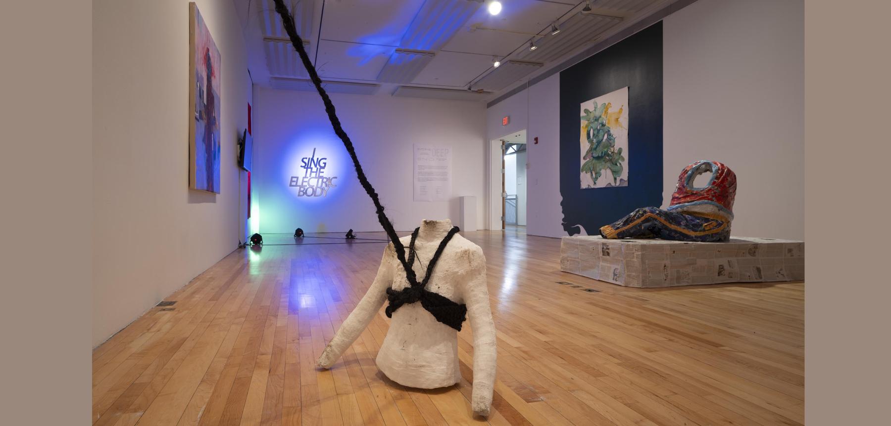 Two torso sculptures placed in a gallery with other paintings on a wall and a lit sign.