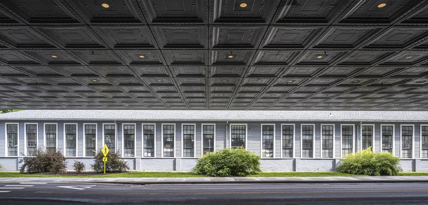 An awning hanging over the street, with a long, white building with lots of windows and several plants across from it.