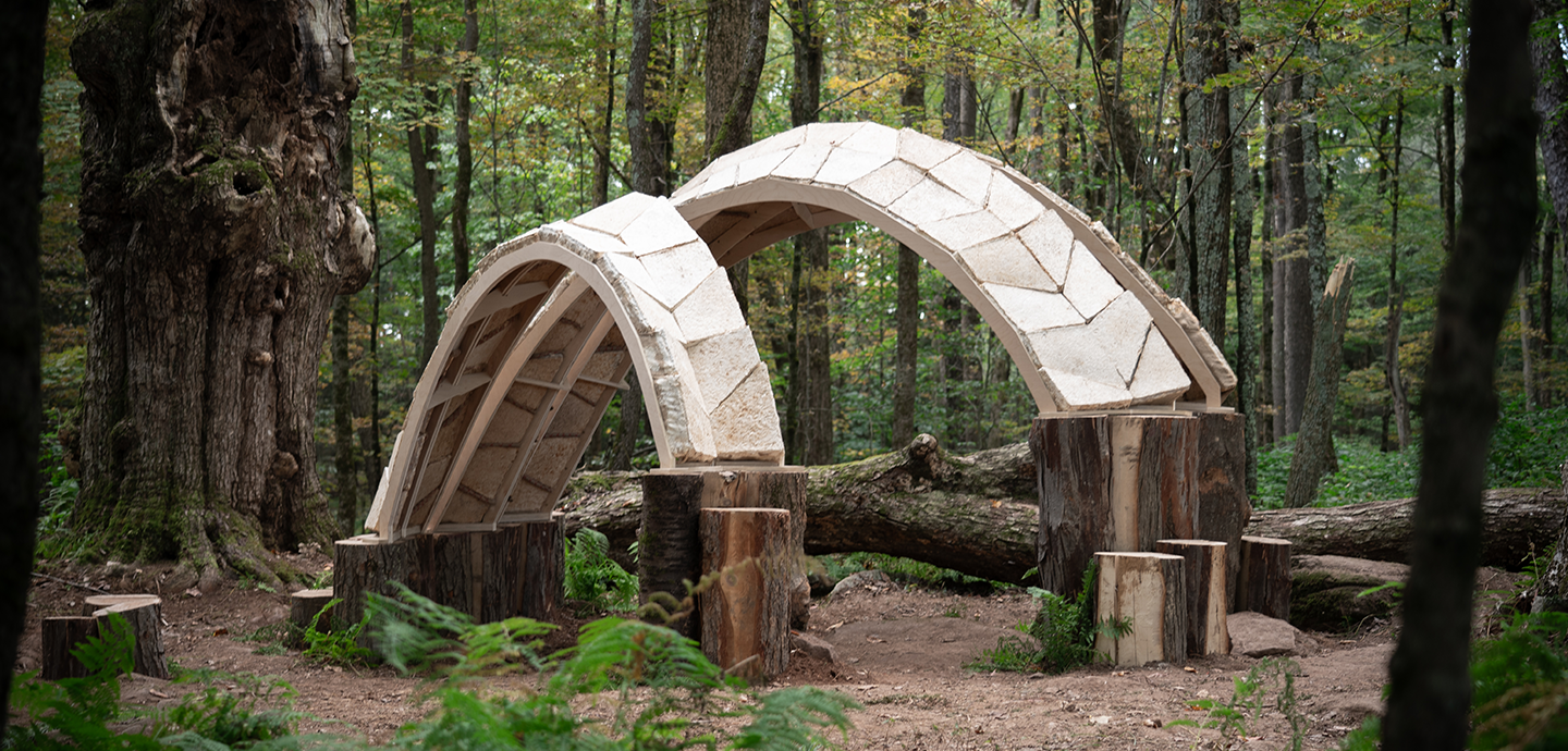 Three arches constructed out of mycelium building blocks are positioned in a forest clearing with stumps used as a foundation.