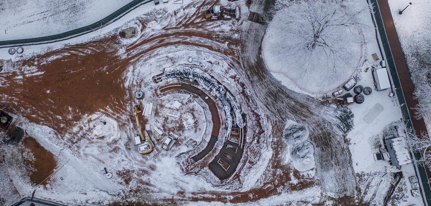 aerial view of construction and site work for a semi circle monument project with a thin layer of white snow on red clay ground