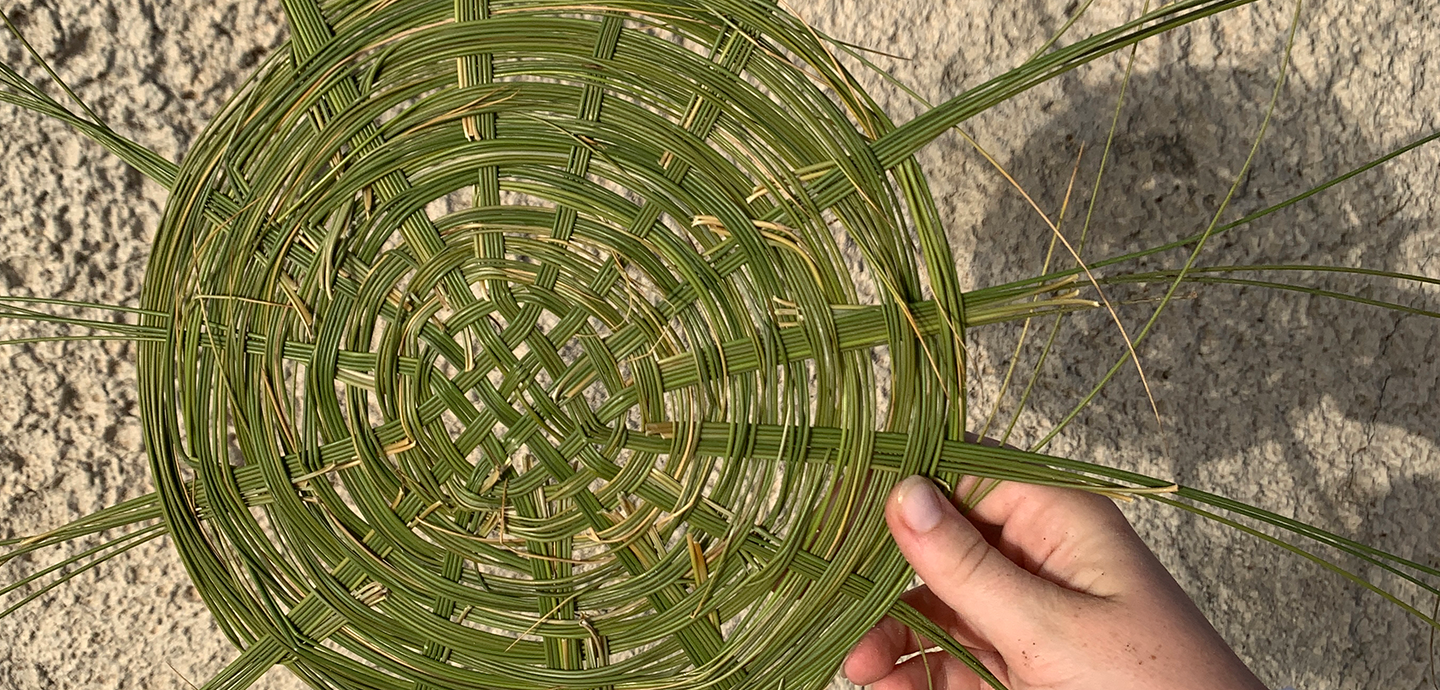 Close-up view of a woven circular grid de out of green grass shoots being held by a pale-skinned hand over a rocky background