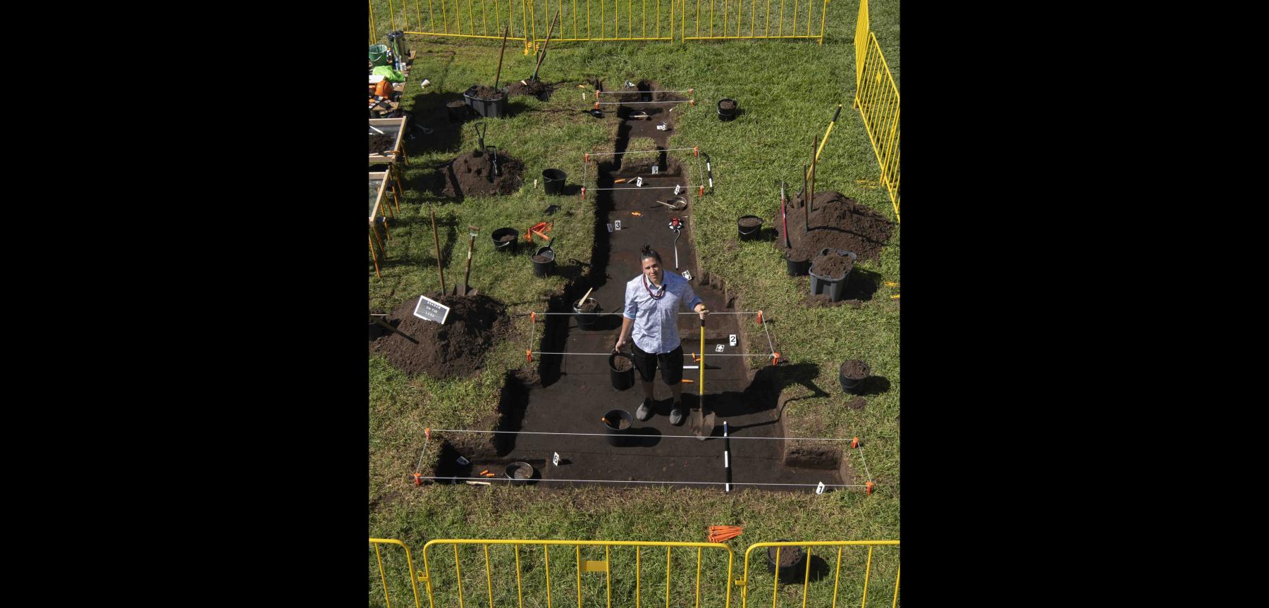 Person wearing a light colored shirt and dark shorts holding a shovel and pail within a dug out sectioned off piece of grass.