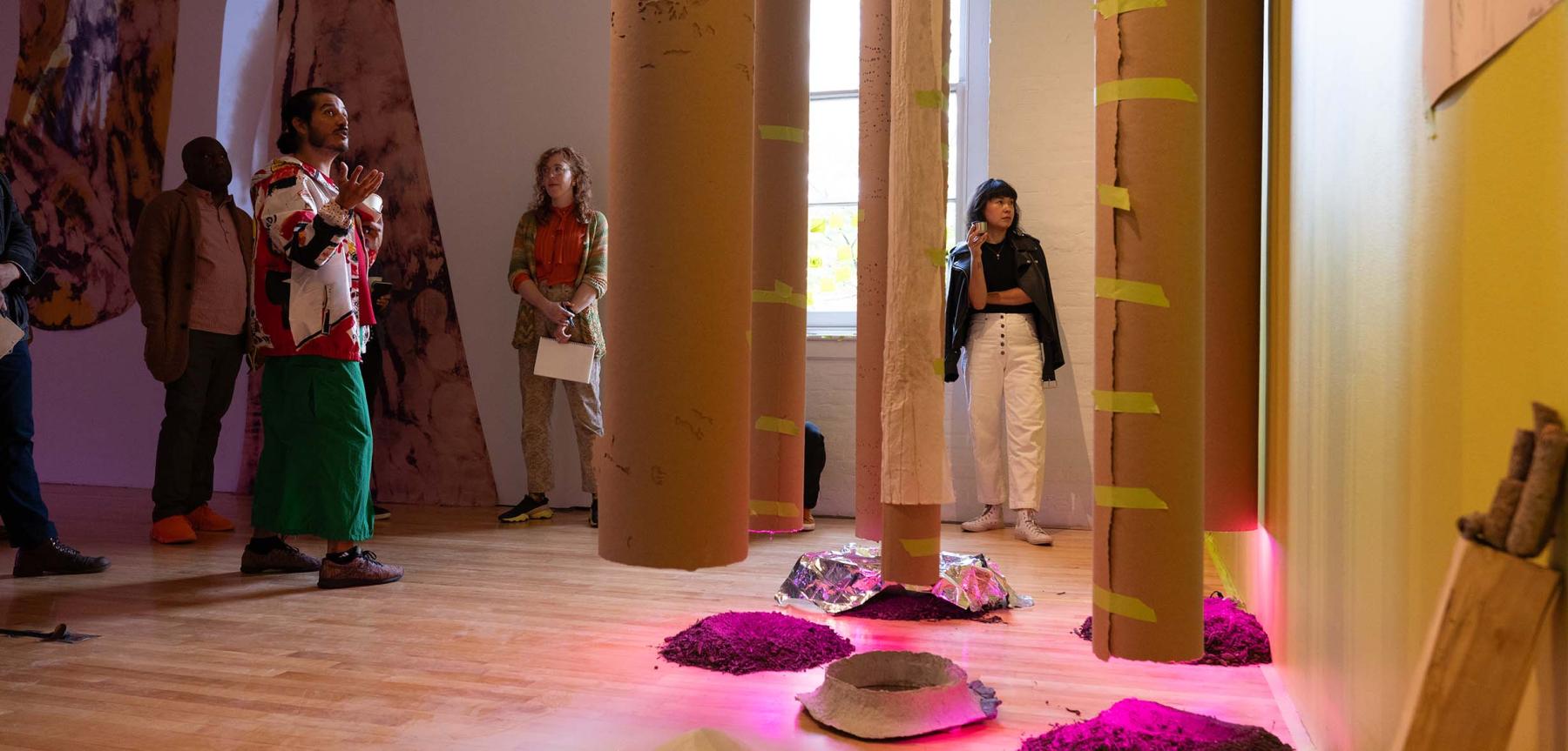 Large paper tubes hanging from the ceiling over piles of dirt on the floor with five people observing.