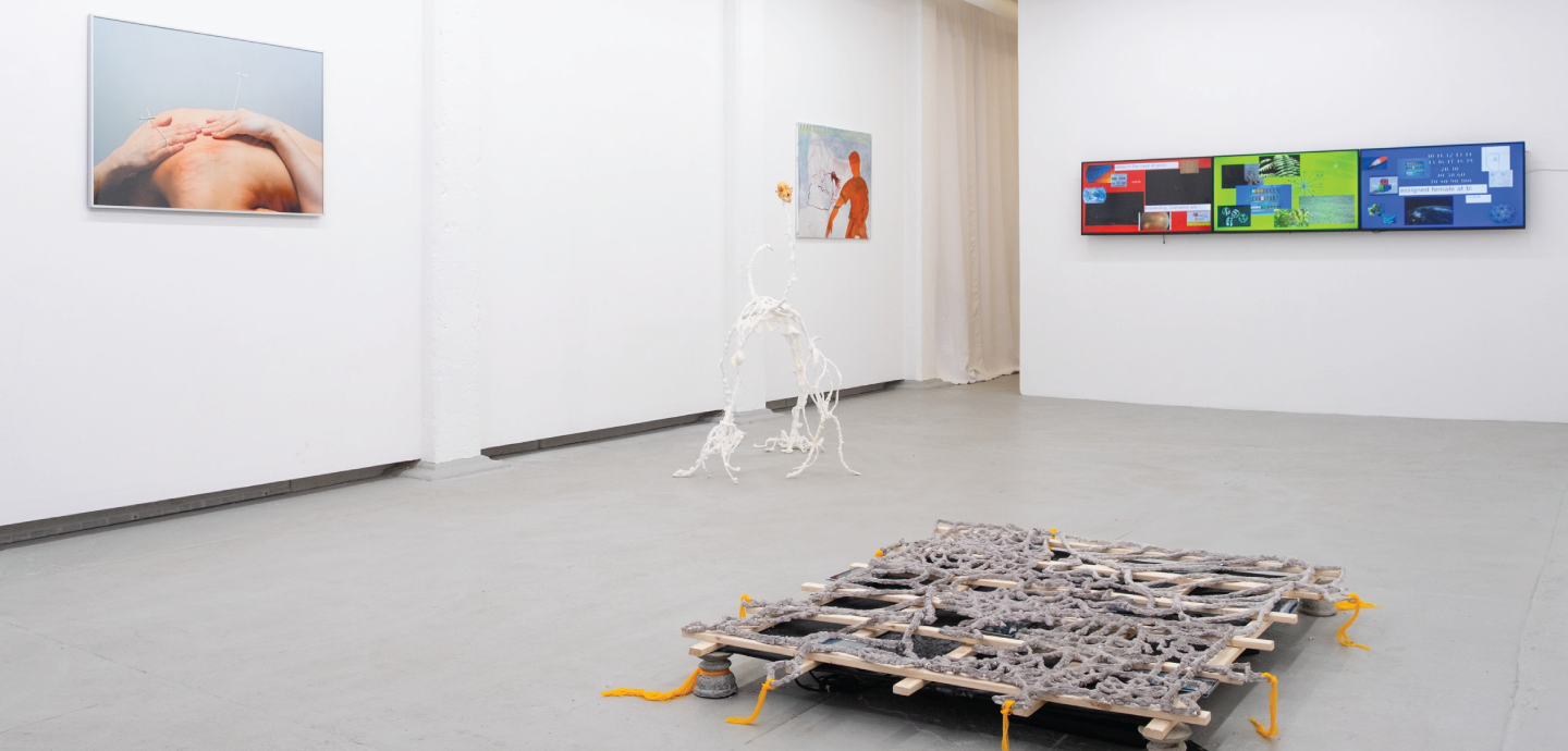 An art gallery with a wooden sculpture in the center, a multi-colored digital panel on the back wall, and two framed images on the left wall.