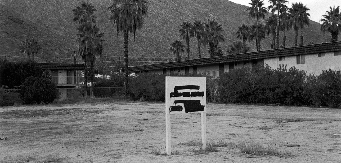 A black and white photo of a white sign with black paint in the middle of a dry field with sparse grass. In the background is a low building, partially obscured by shrubs, with palm trees and a distant hill rising up in the back.