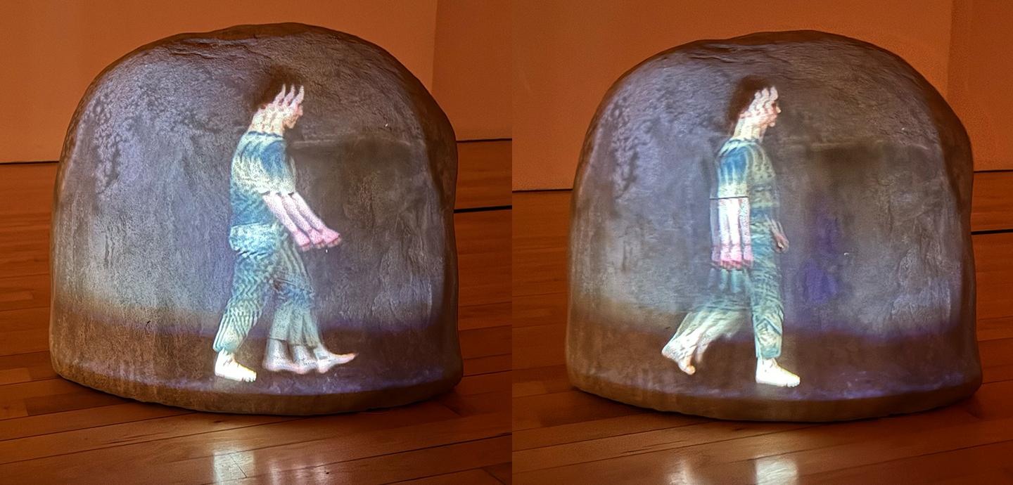 A large rock on a wooden floor, with a projected image of a person with brown hair and matching blue and green shirt and pants, mid-walk. There are multiple, overlapping projections, leading to a slightly distorted image,