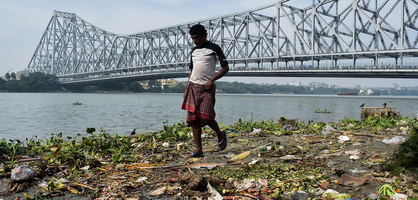 Man wearing a tshirt and sarongs walks on a river bank littered with garbage with a large modern bridge in the background