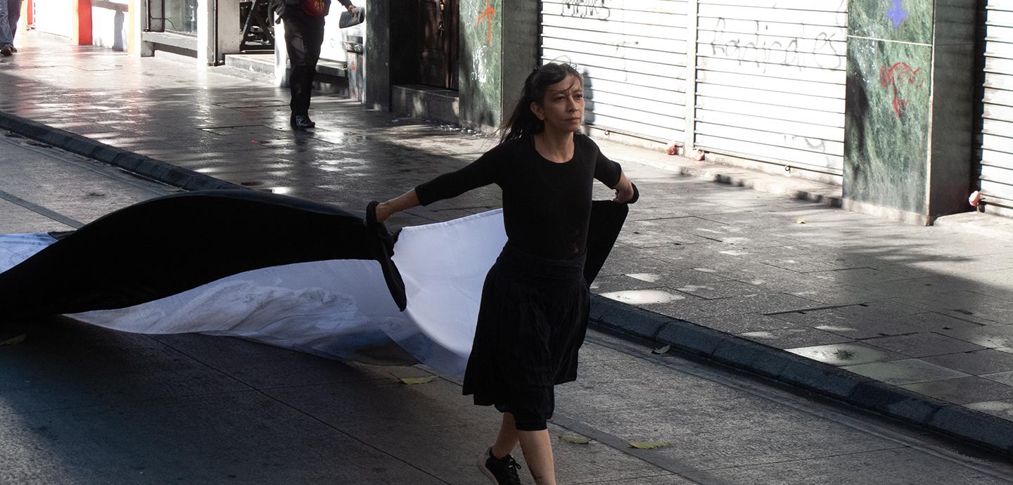 A petite woman with dark hair and wearing a black dress passes a graffitied building as she walks down a street, holding a large black and white flag behind her.