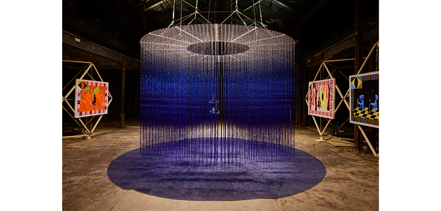 Circular sculpture installation made out of black and blue string or textile hangs in the center of a room, surrounded by paintings in diamond-shaped wooded frames. 
