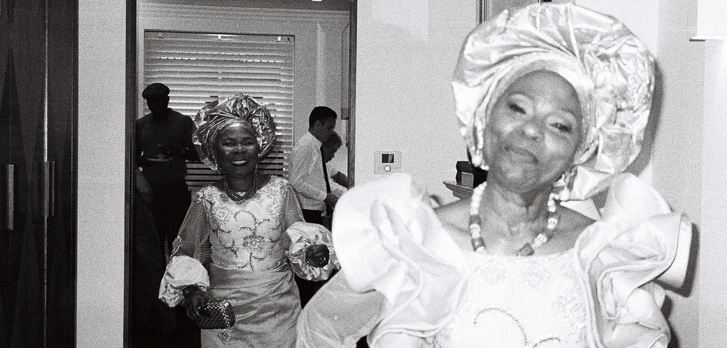 A black and white photo of a woman wearing a dress with lots of intricate details and a head scarf as she looks at the camera. Behind her is another woman who is looking at her and smiling.