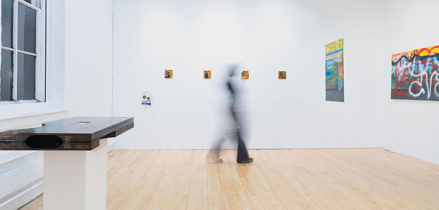 An art gallery with a blurred person walking past in the center, colorful prints on the walls, and a large wooden sculpture on a pedestal.