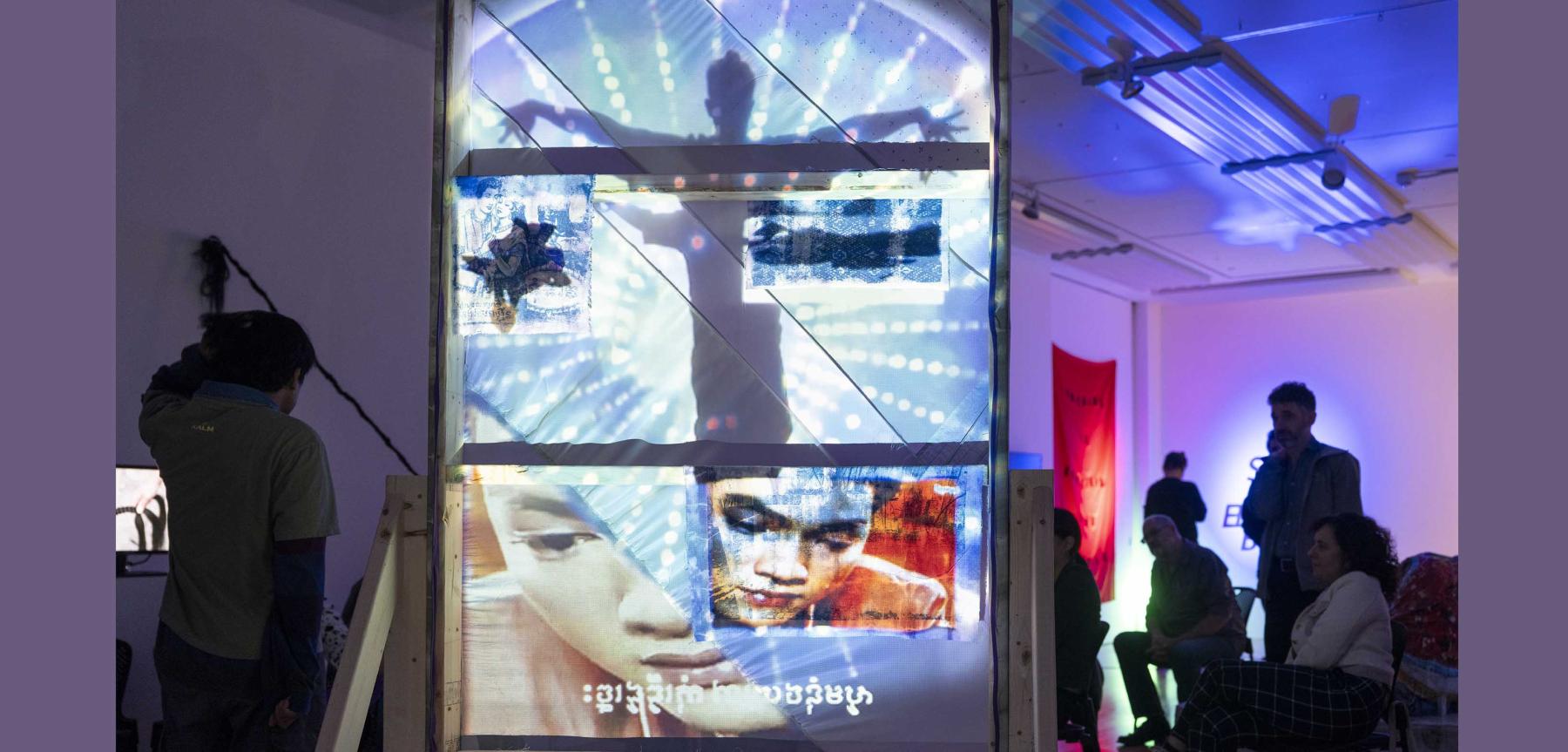 Dark purple lit space with people looking at a video projection on a tall wooden structure.