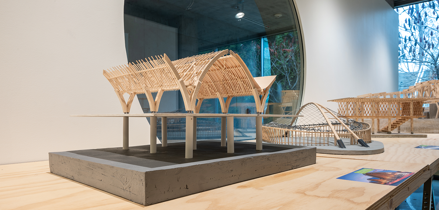 Three wood and concrete architectural models on plywood tables with a porthole window in the background.