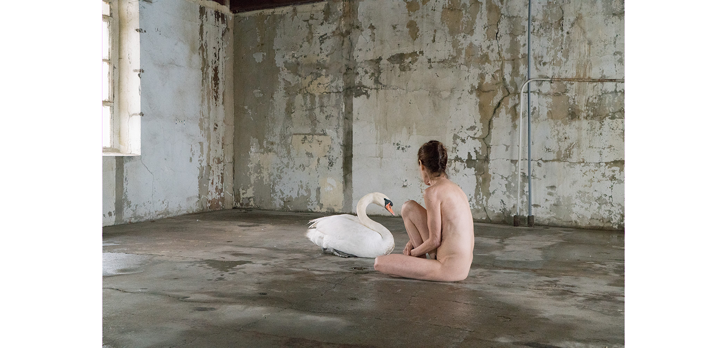 A naked person with pale skin sits next to a white swan on the floor of a concrete room.