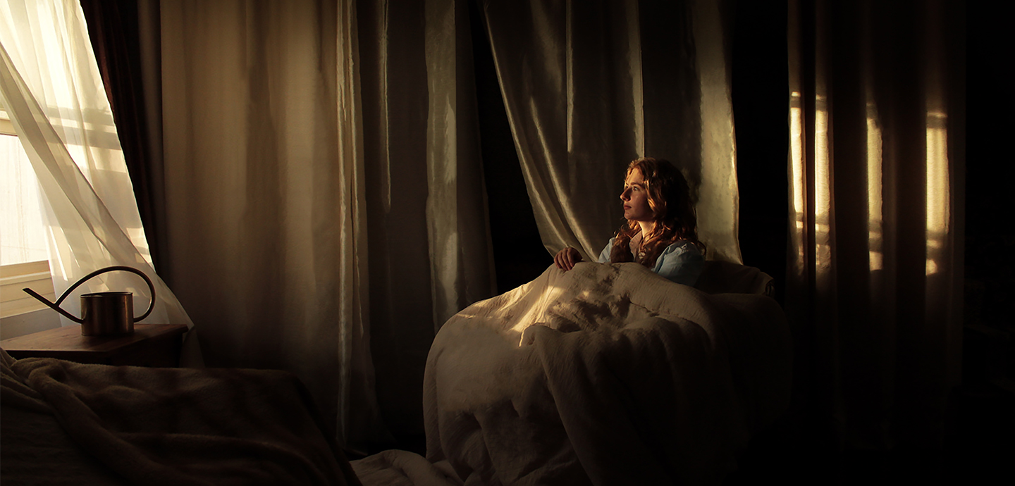 Person with long blonde hair sits in a chair covered by a white blanket and stares out a window loosely covered by sheer drape.