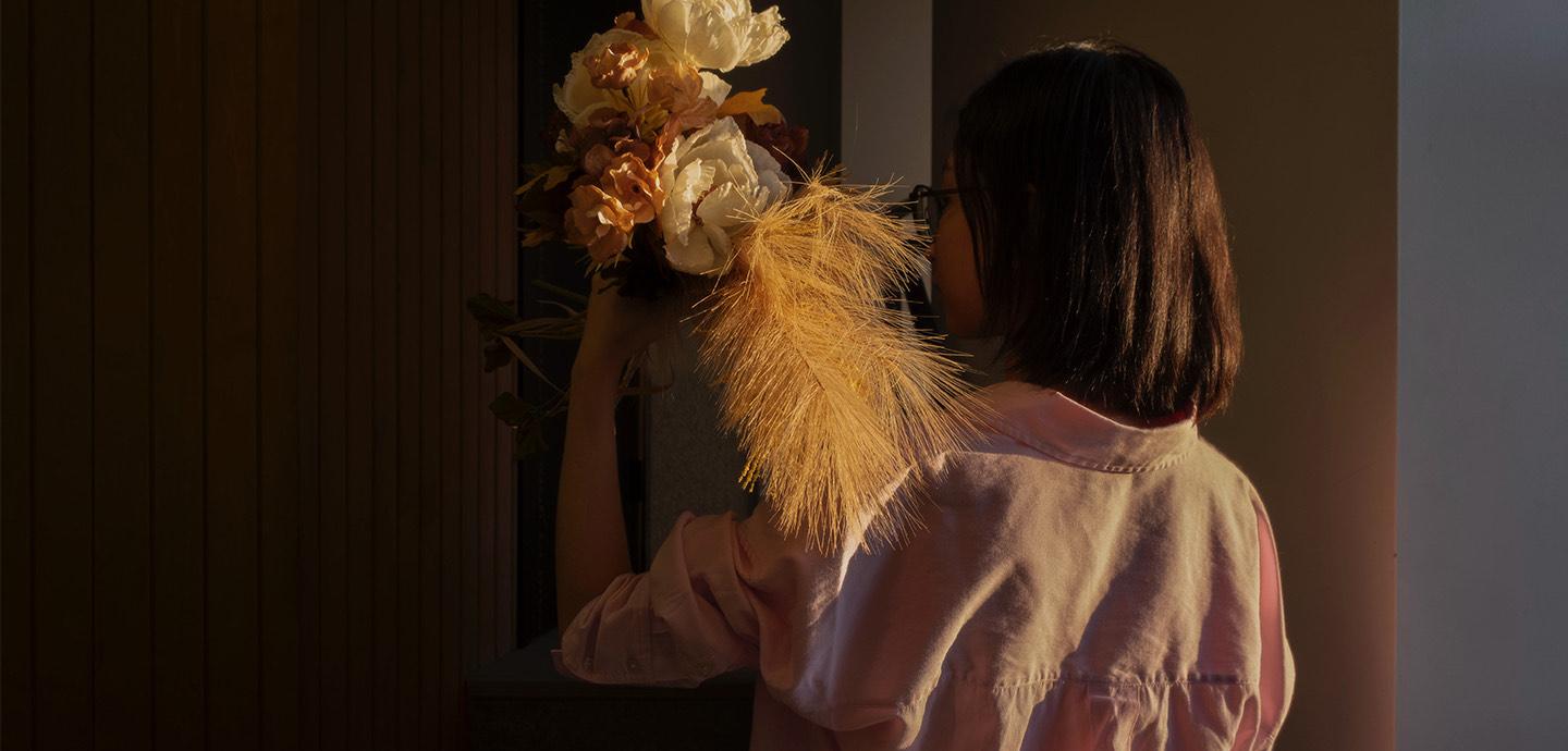 Person holding a bouquet of flowers next to their face with their body turned to the wall.