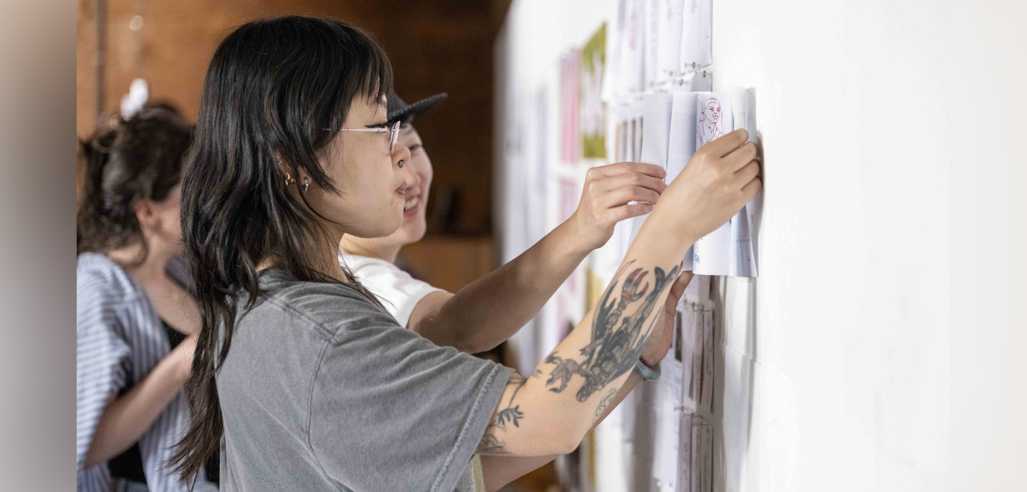 Person with long dark hair in a grey shirt with tattoos hanging papers on a white wall.