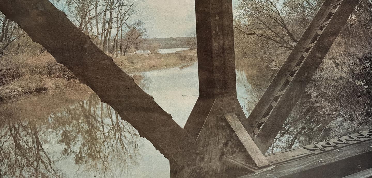 A muted color photograph of the steel beams of a bridge. Seen through the gaps is a lake with barren trees lining the banks,