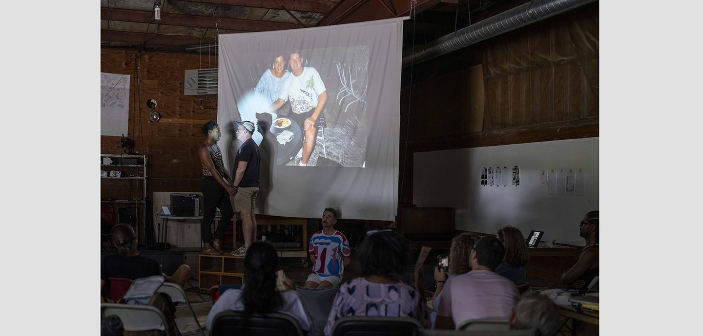Three people performing on a small stage in front of a projected picture in front of a small audience.
