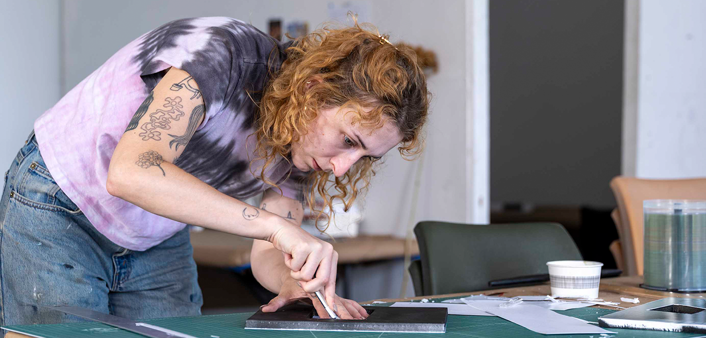 Woman with dark blonde curly hair cutting a black piece of paper on a green cutting board.