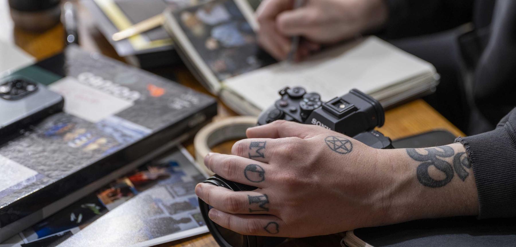 Hand with small black tatoos holding a camera surrounded by books and notebooks on a desk.