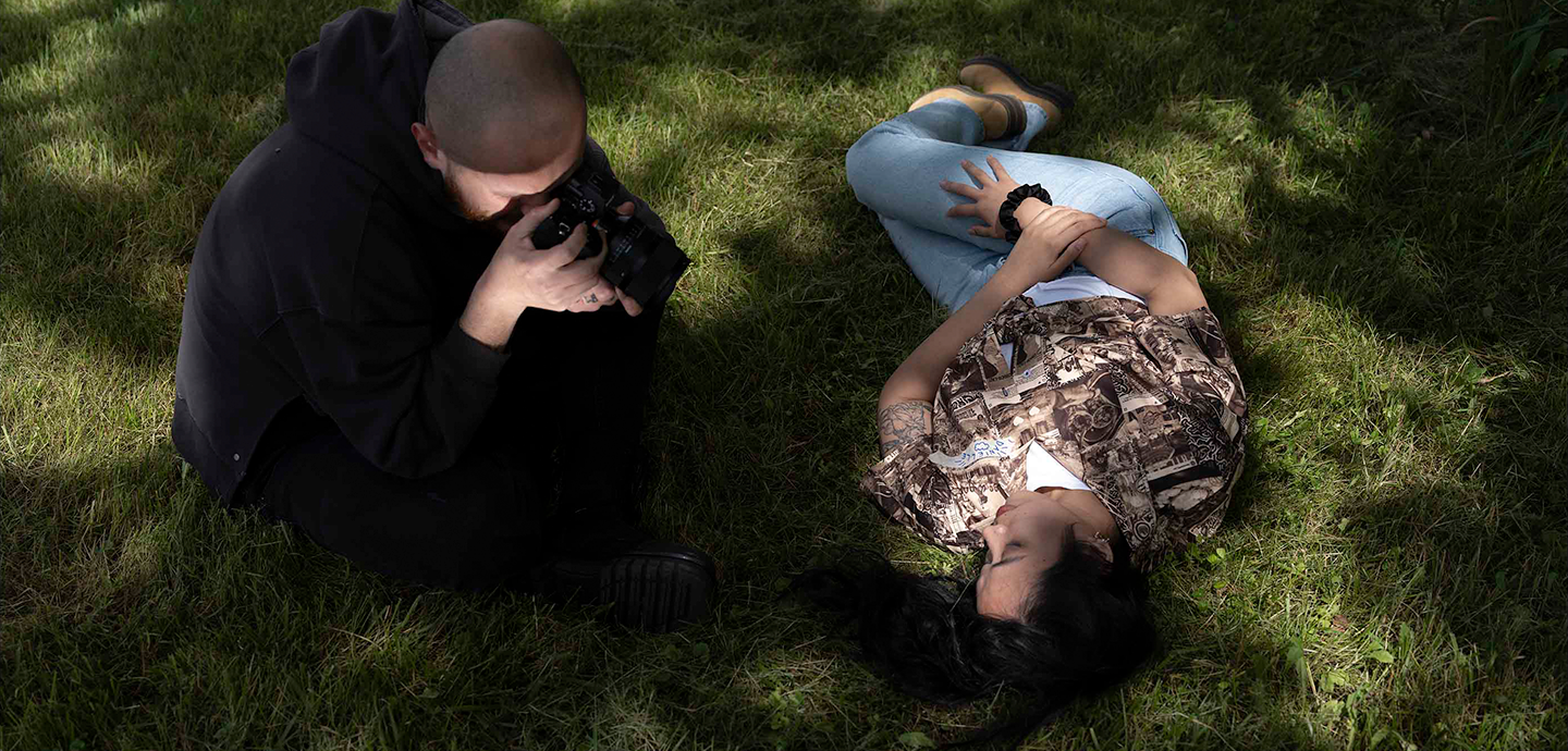 Two people on shadowed green grass, one laying down, while the other takes their picture with a circular mirror angled up.