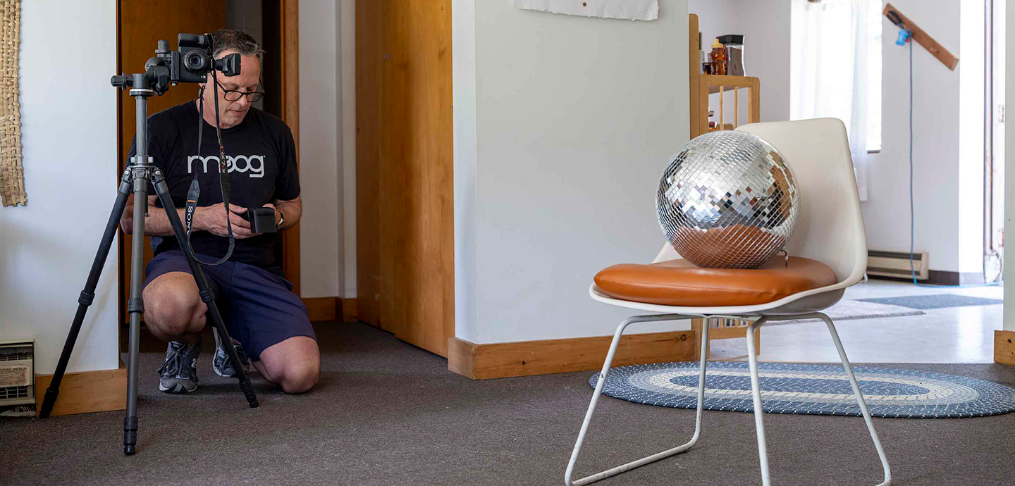 Person kneeling down to take a photograph of a disco ball on a small white chair.