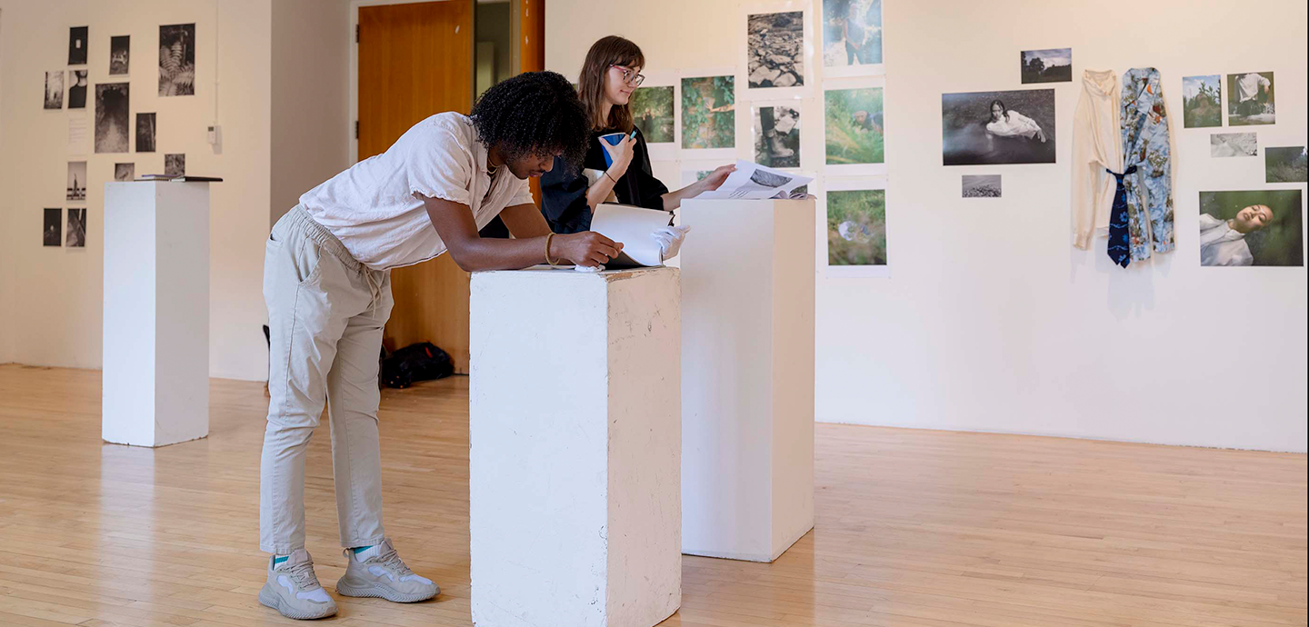 Two people examining artwork on pedestals in a gallery with various pictures on the wall.