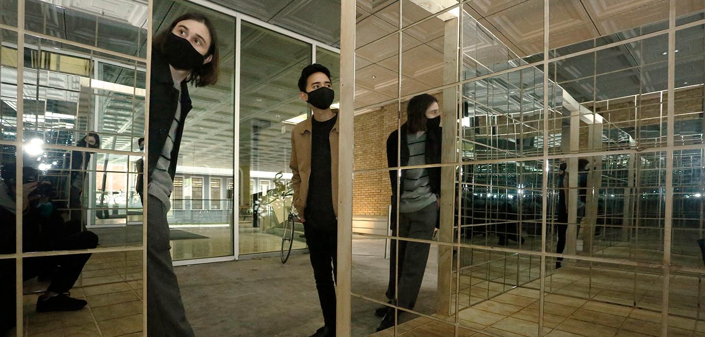 Two people with masks in the doorway of a mirrored box structure.