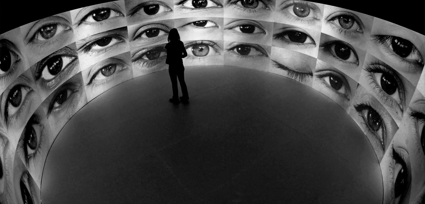 A person standing in the middle of a circular room with walls completely covered by a projection of multiple eyes.