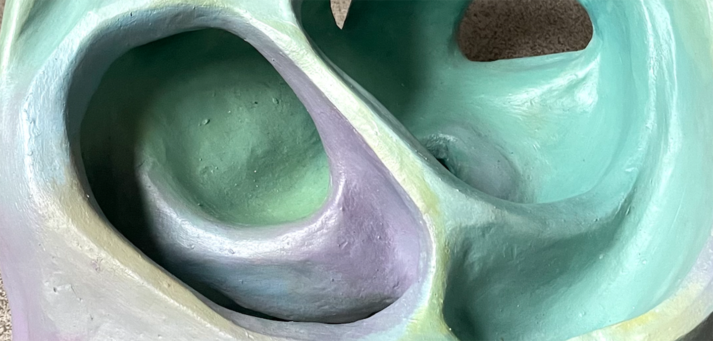 A round clay sculpture with many large holes, painted in shades of blue, green and purple