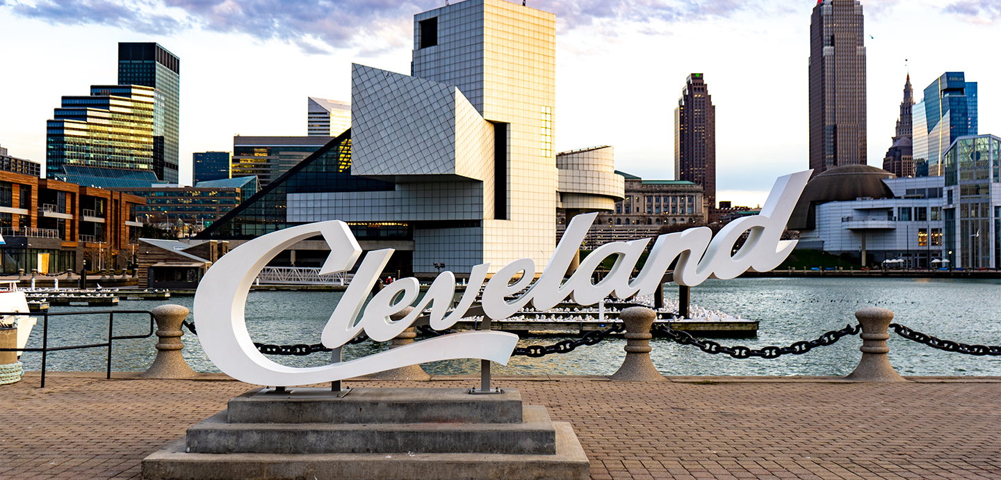City landscape with a sign of of Cleveland with buildings and a body of water in the background.