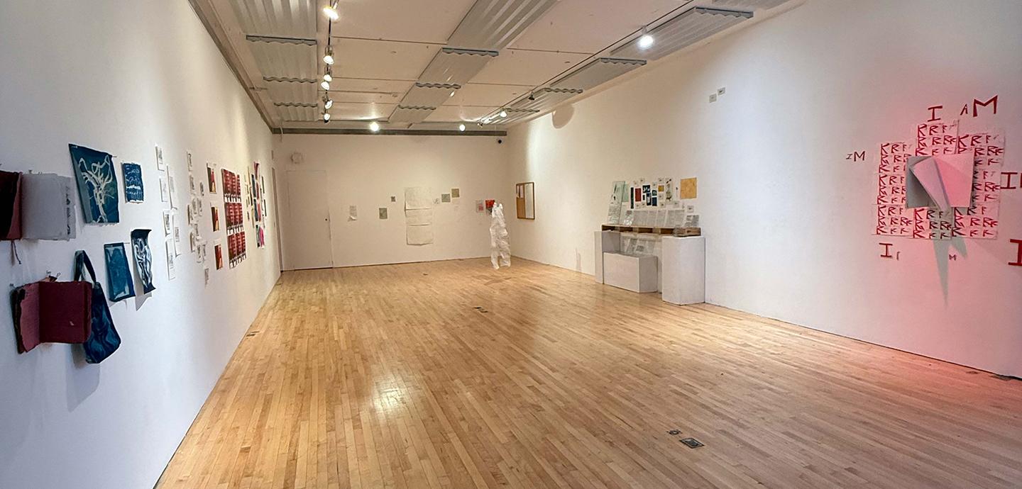 An installation view of an art gallery: mounted on the right wall is a book with red text, the left wall has four blue and white prints, and there are more indistinct art works in the background. 