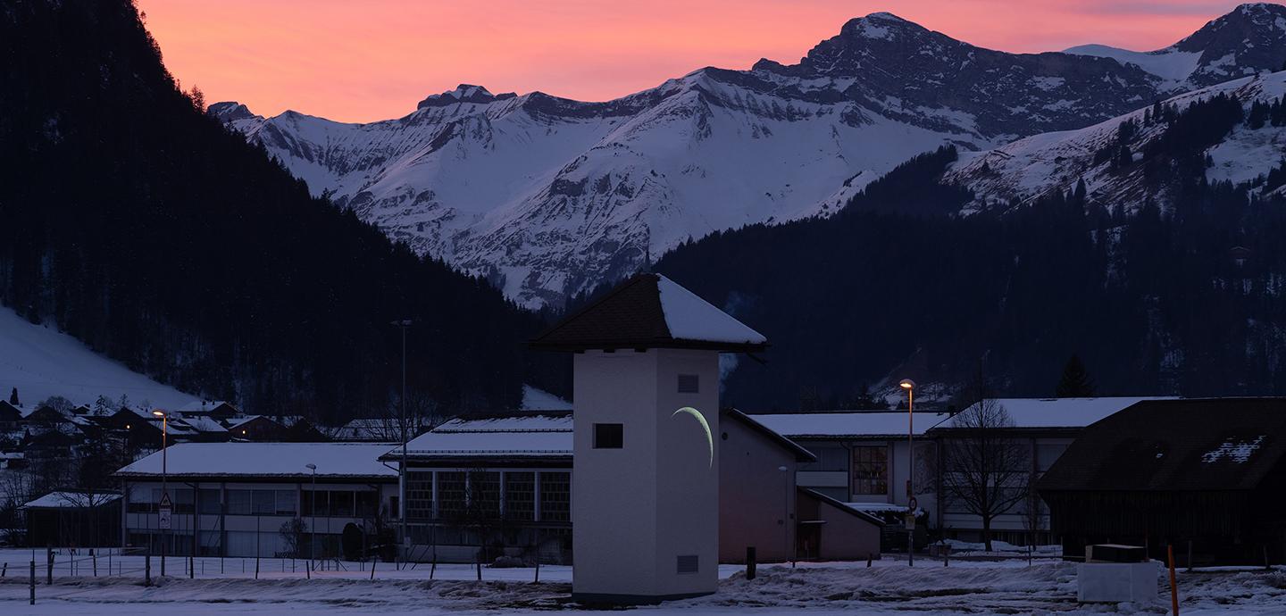 A snowy landscape at sunset, with a long, narrow building in the foreground and mountains in the background